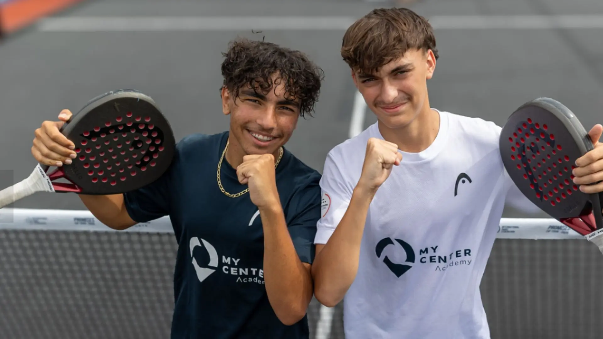 Two young men on a padel court pose with fists up, holding rackets, wearing My Center Academy shirts and smiling at the camera.