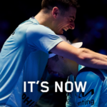 Two athletes in light-blue jerseys embrace in a dark arena, with bold white text 'IT'S NOW' at the bottom.
