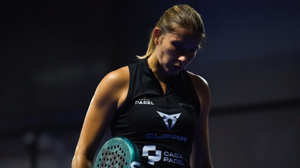 Female padel player in a black sleeveless top holding a teal paddle, looking down against a dark background.