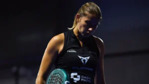 Female padel player in a black sleeveless top holding a teal paddle, looking down against a dark background.
