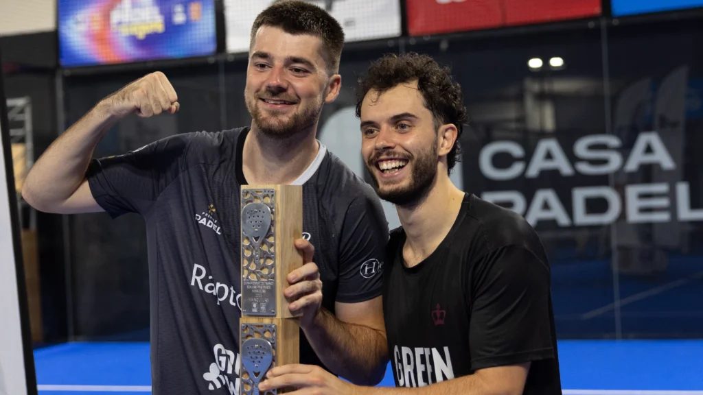 Two smiling men celebrate, posing with a tall wooden trophy in a sports facility backdrop.