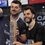 Two smiling men celebrate, posing with a tall wooden trophy in a sports facility backdrop.