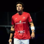 Male padel player in a red jersey standing on a court with a paddle in hand, looking up, dark background behind him.