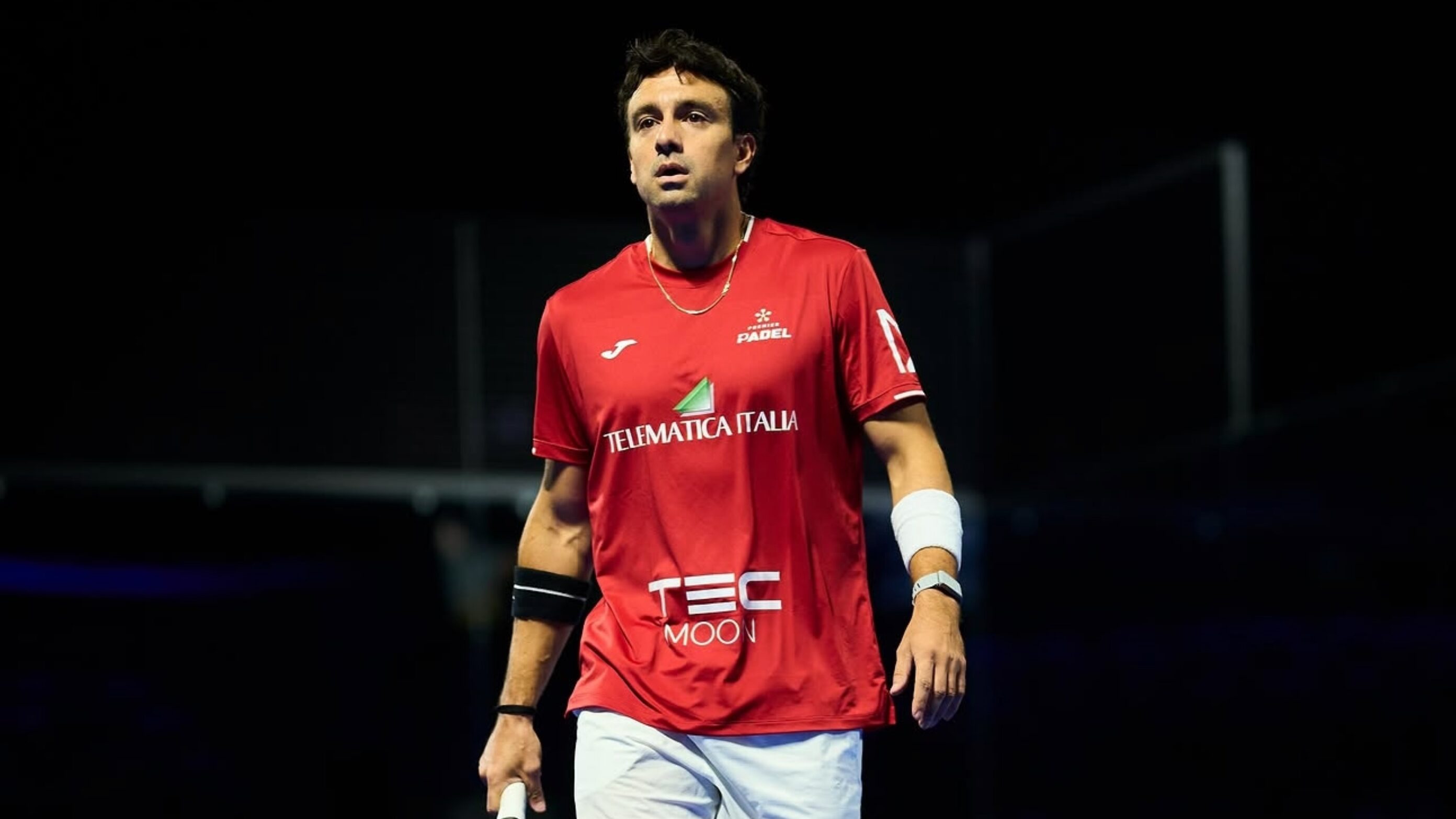 Male padel player in a red jersey standing on a court with a paddle in hand, looking up, dark background behind him.
