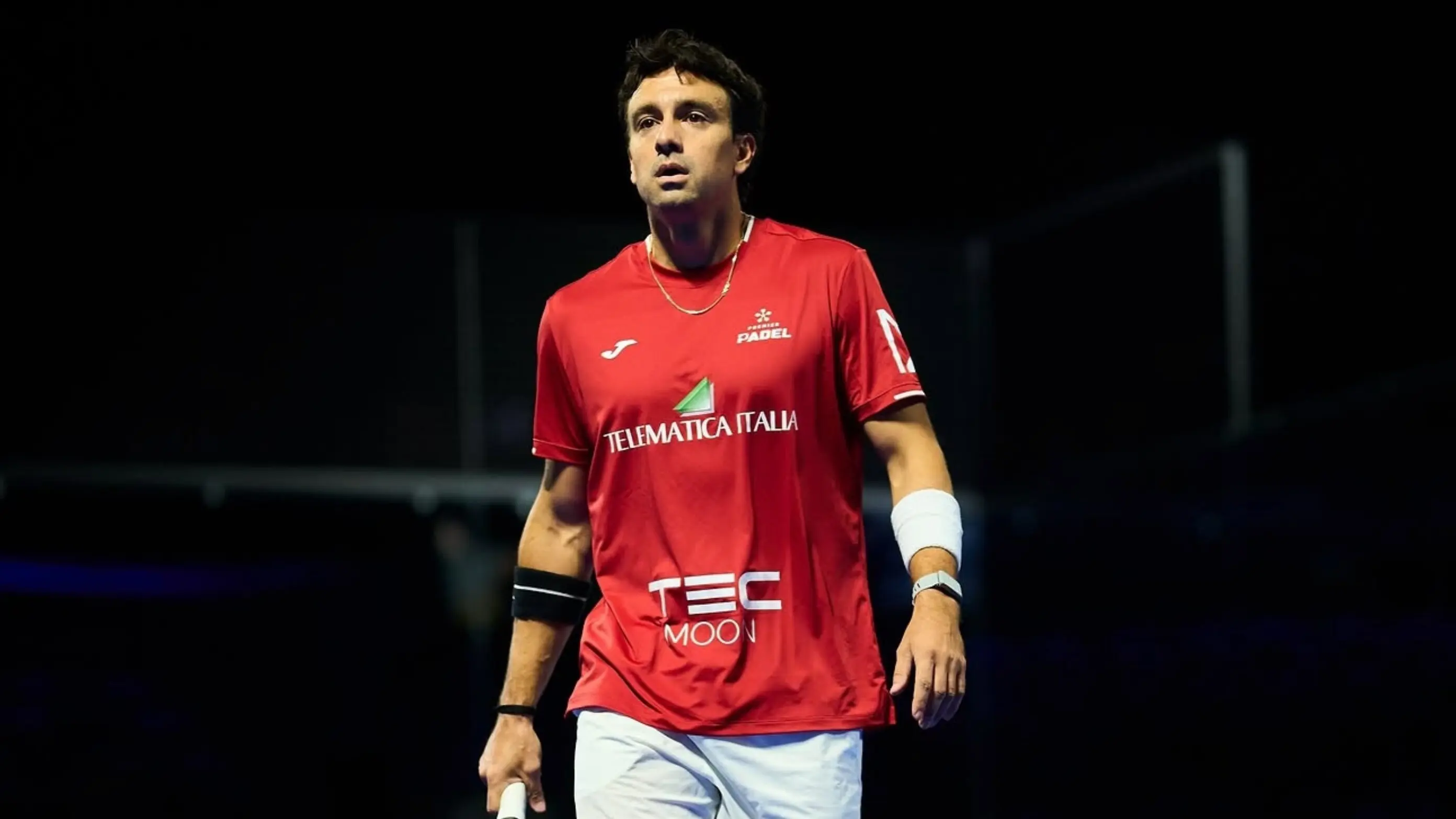 Male padel player in a red jersey standing on a court with a paddle in hand, looking up, dark background behind him.