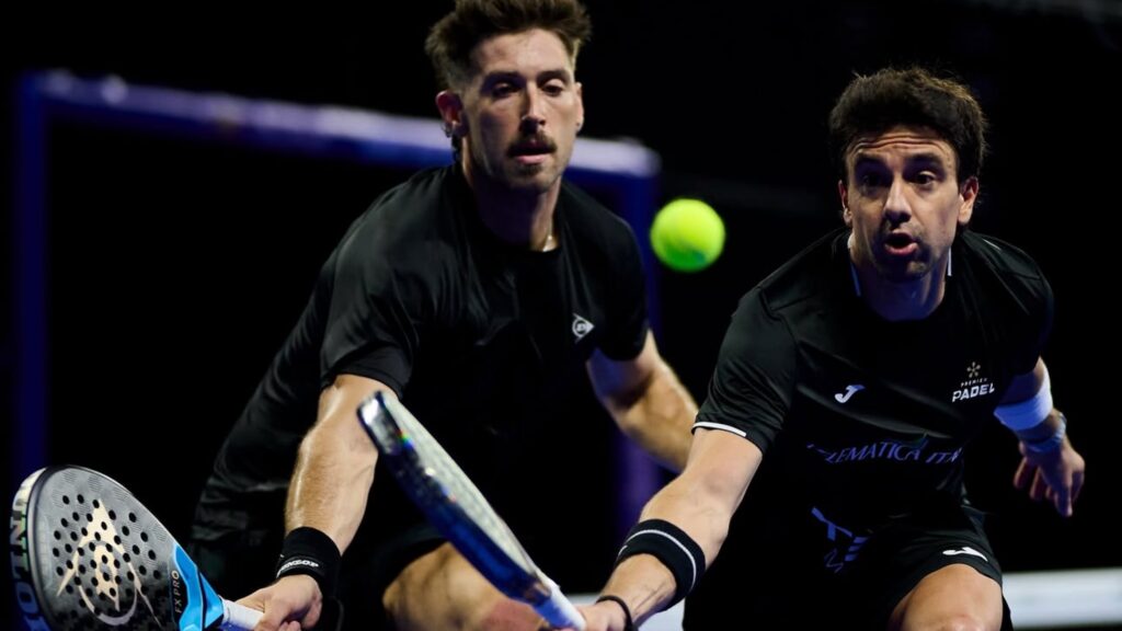 Two male padel players in black shirts reach for a bright yellow ball during an intense match on an indoor court.