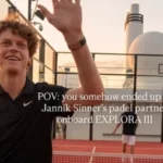 Smiling man in a black polo waves at the camera on a rooftop tennis court; caption mentions paddle partner Janik Sinner onboard EXPLORE III.