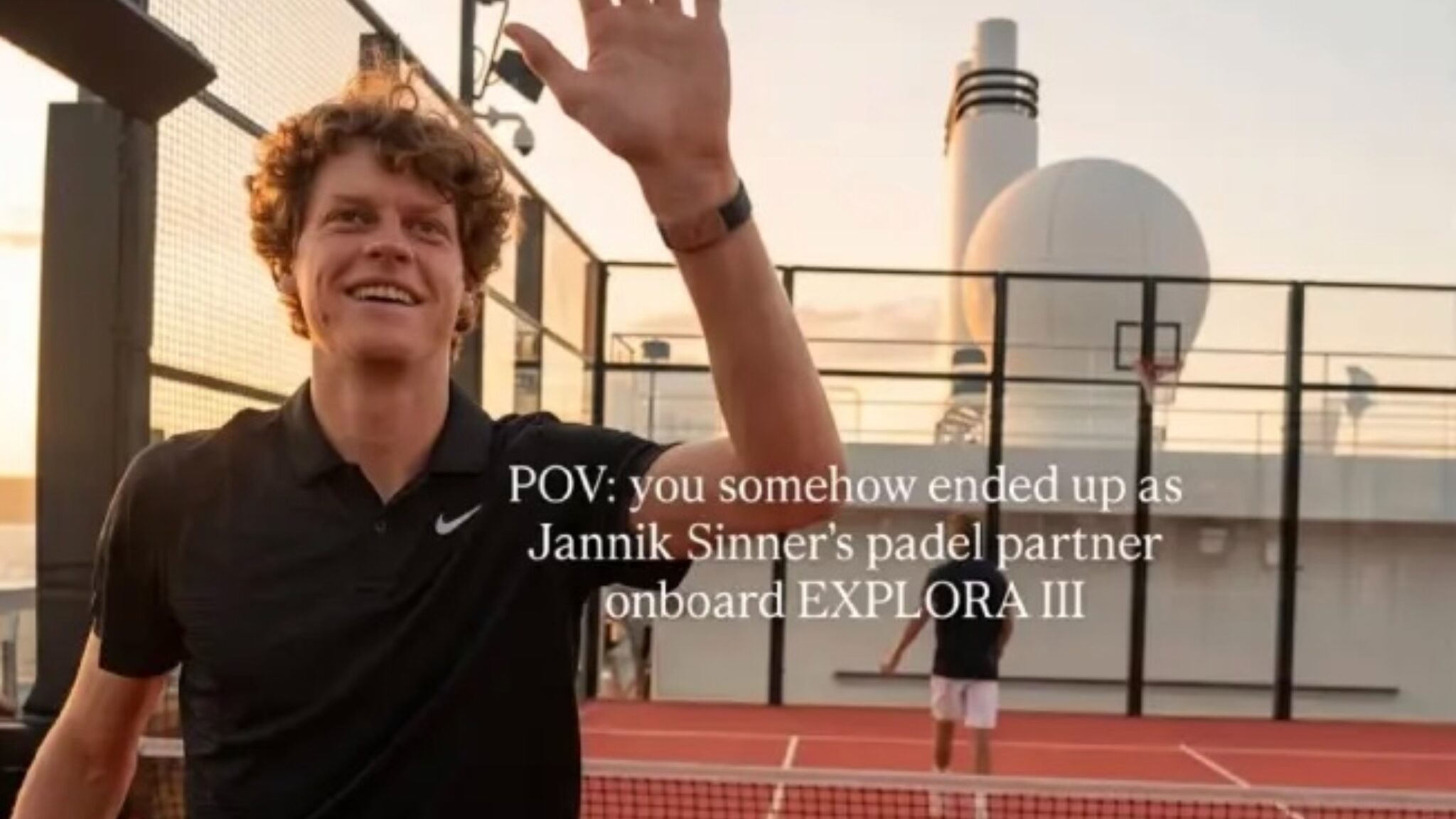 Smiling man in a black polo waves at the camera on a rooftop tennis court; caption mentions paddle partner Janik Sinner onboard EXPLORE III.