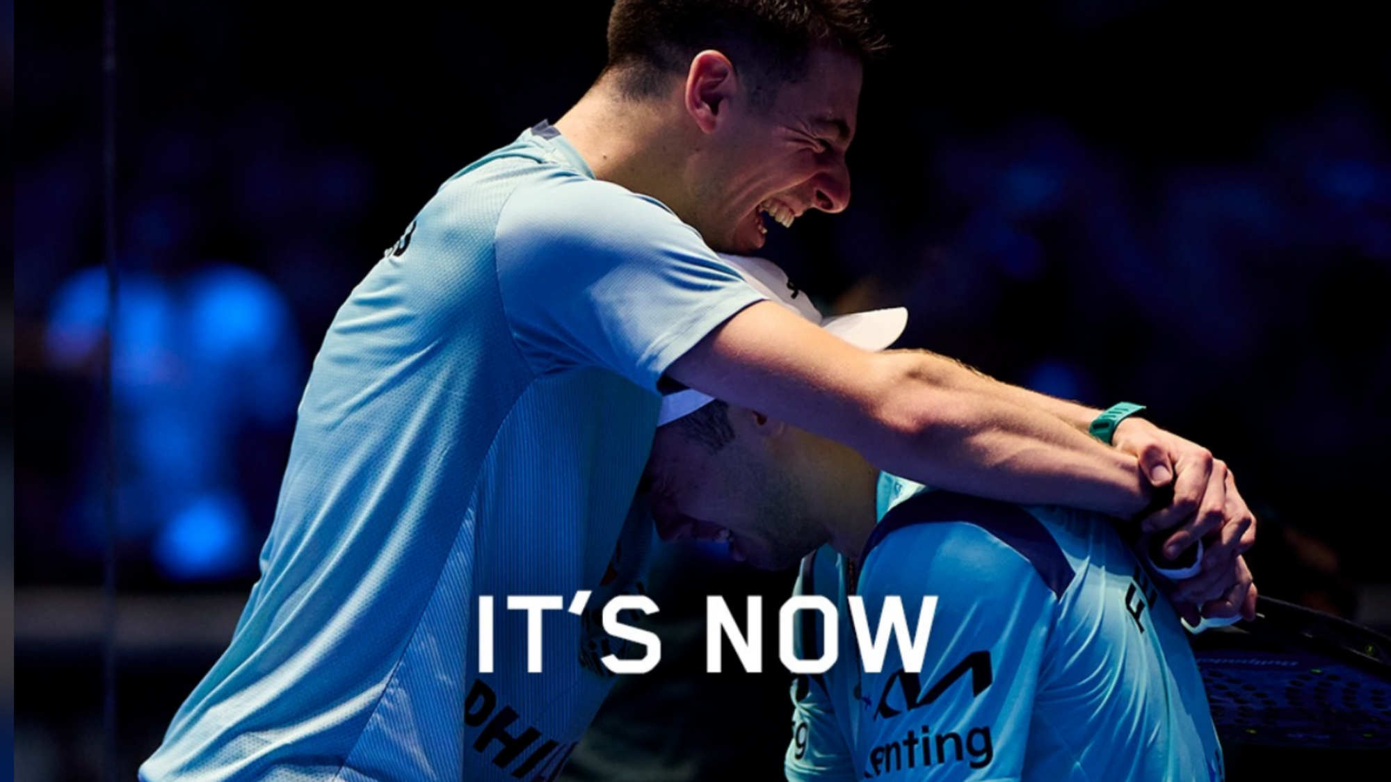 Two athletes in light-blue jerseys embrace in a dark arena, with bold white text 'IT'S NOW' at the bottom.