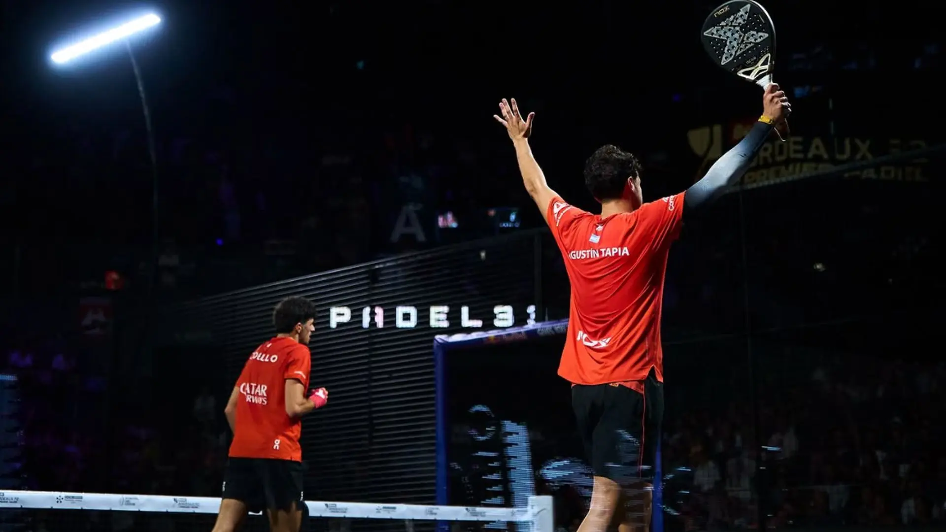 Celebration moment in a padel match: a player in red raises a racquet overhead on a dark arena backdrop