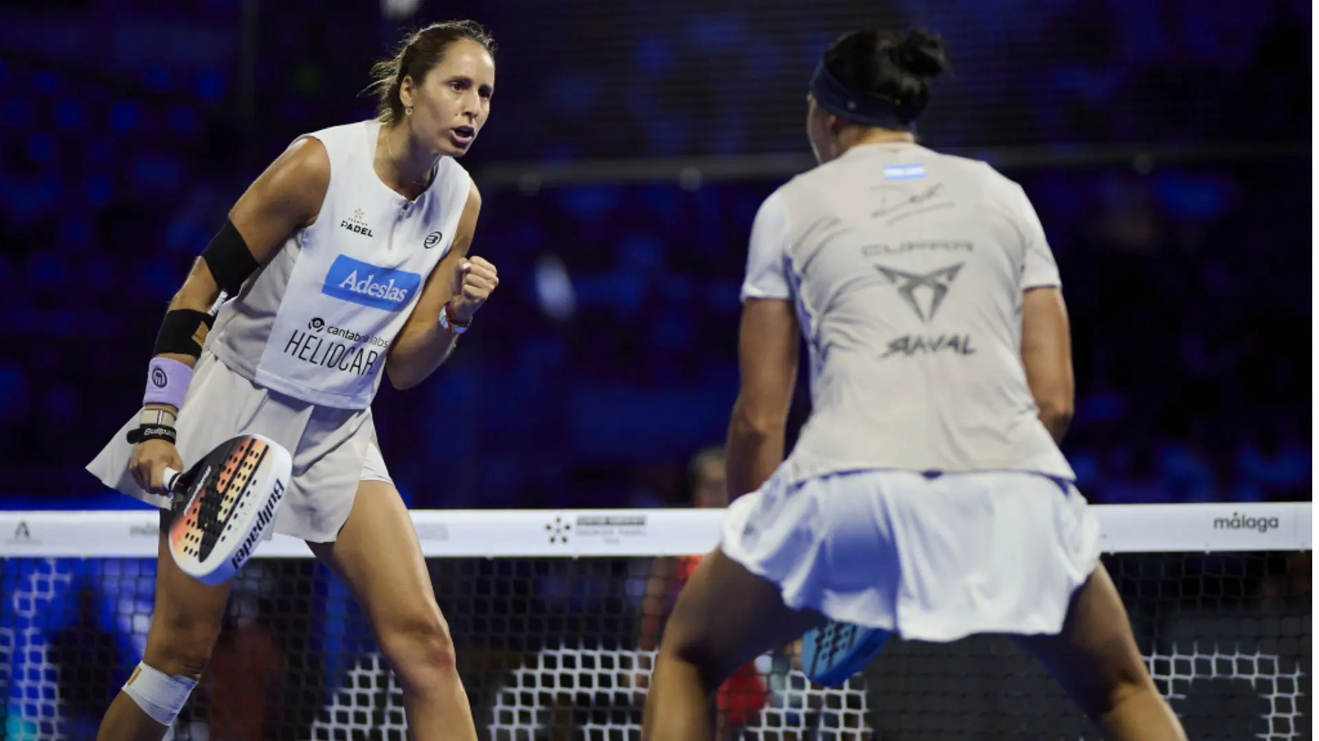 Two female padel players near the net during a match, one preparing to hit the ball with her paddle as the other watches from the opposite side of the court.