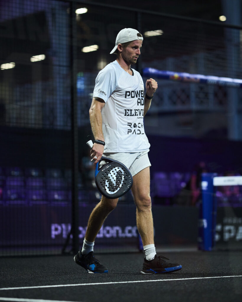 Leygue / Geens renversent Cepero / Torre : un exploit majuscule qui envoie un message fort | Padel Magazine Male padel player in a white shirt and cap on an indoor court, fist raised in a triumph pose, holding a perforated paddle.