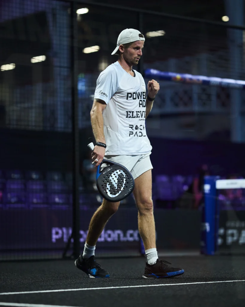 Jugador de pádel masculino con camiseta y gorra blancas en una pista cubierta, con el puño en alto en señal de triunfo, sosteniendo una pala perforada.