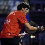 Male athlete in an orange shirt grips a squash racket during a match, focused on the play.