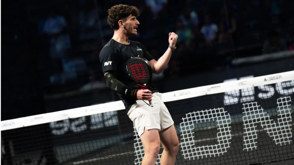 Male padel player in a black shirt and white shorts raises a clenched fist in celebration at the net during a match.
