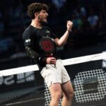 Male padel player in a black shirt and white shorts raises a clenched fist in celebration at the net during a match.