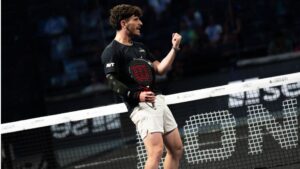 Male padel player in a black shirt and white shorts raises a clenched fist in celebration at the net during a match.