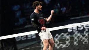 Male padel player in a black shirt and white shorts raises a clenched fist in celebration at the net during a match.