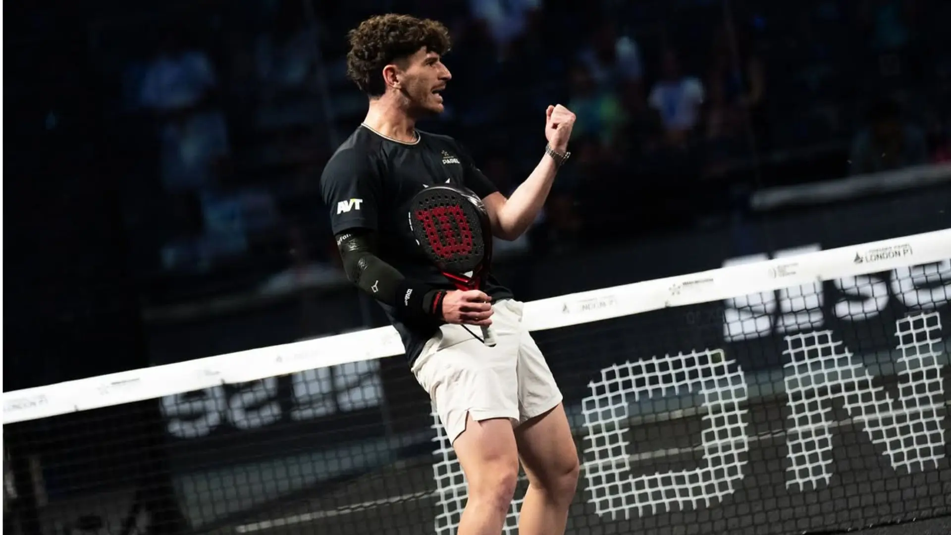 Male padel player in a black shirt and white shorts raises a clenched fist in celebration at the net during a match.