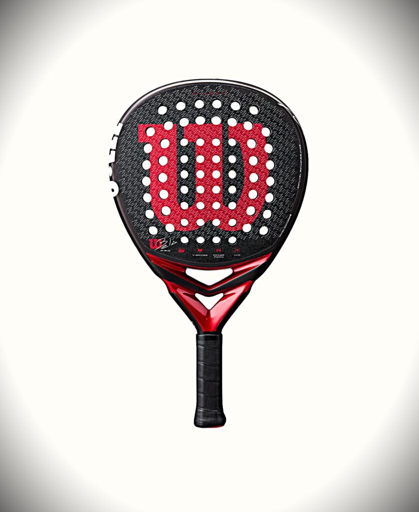 Bela Pro | Padel Magazine Padel racket with black textured face, red emblem pattern, numerous white holes, and a red-and-black grip grip at the bottom.