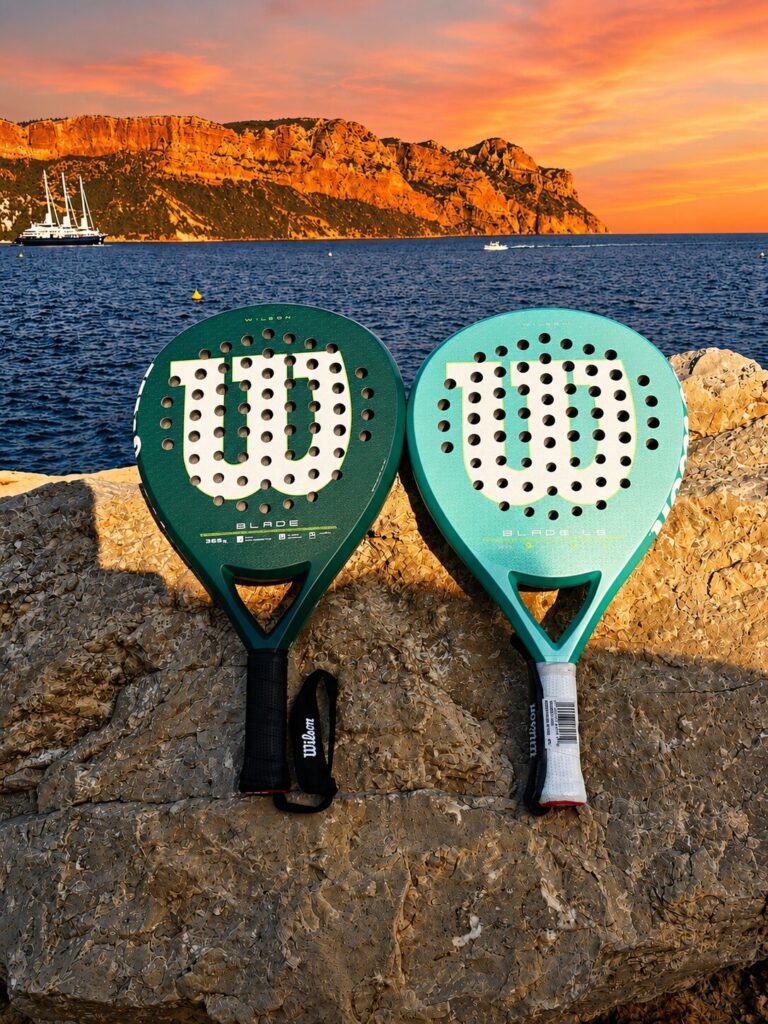 Blade Cassis | Padel Magazine Two paddle tennis rackets rest on a rocky ledge by the sea at sunset, with orange cliffs in the background and a boat on the water behind them.