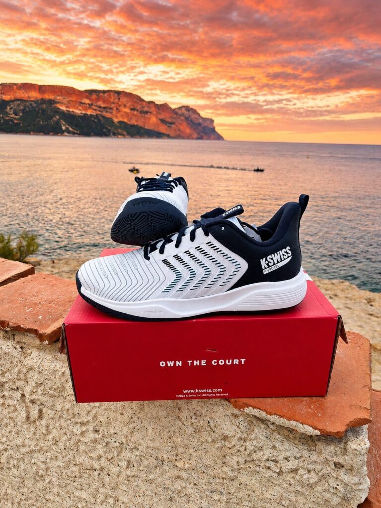 Cassis 1 | Padel Magazine White and black K-Swiss running shoes resting on a red box by a rocky wall at sunset over the sea.