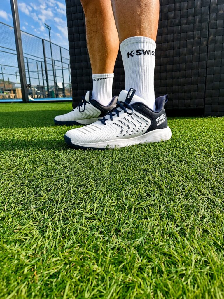 Chaussures herbe | Padel Magazine Close-up of white and navy running shoes on artificial grass, worn with white KS WISS socks beside a wicker outdoor chair or couch area in a sports setting.