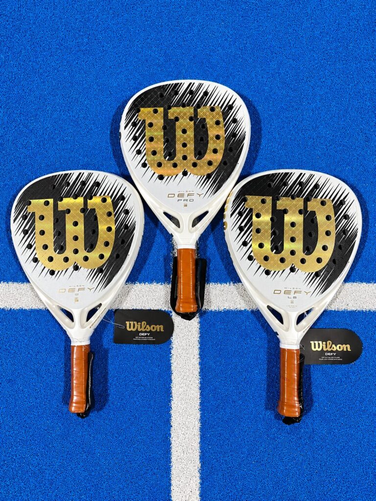 Three Wilson pickleball paddles with gold branding arranged on a blue court with a white line, showing a gear setup for pickleball equipment