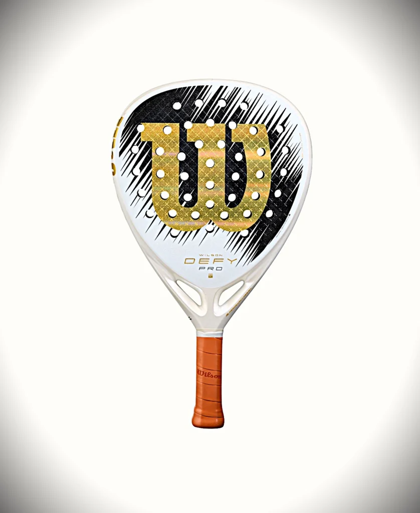 Defy V1 Pro | Padel Magazine 白いウィルソン製パデルラケット。金色の7番のロゴ、黒いストライプの背景、オレンジ色のグリップハンドルが特徴です。