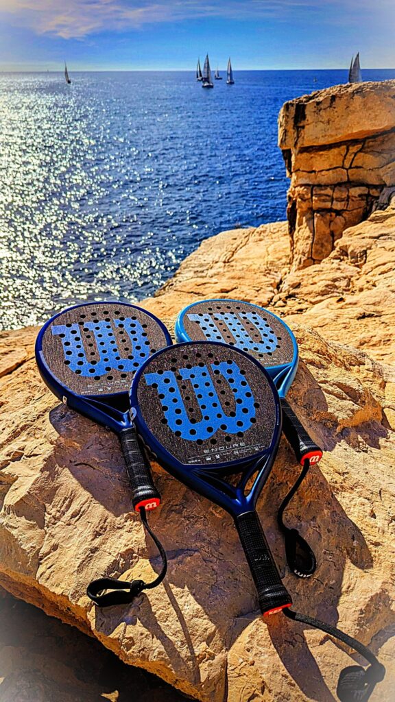 Three blue perforated paddle racquets resting on a rocky cliff by the sea with sailboats in the distance