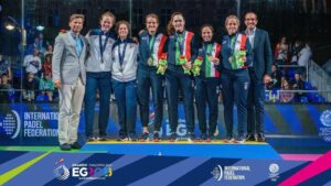 Eight athletes stand on a podium with medals, flanked by two coaches, at a padel event.