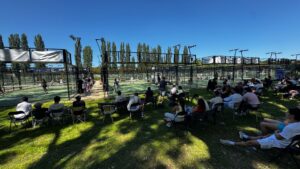 Crowd watching an outdoor padel match on fenced courts under a bright blue sky, with players in action and spectators on benches nearby.