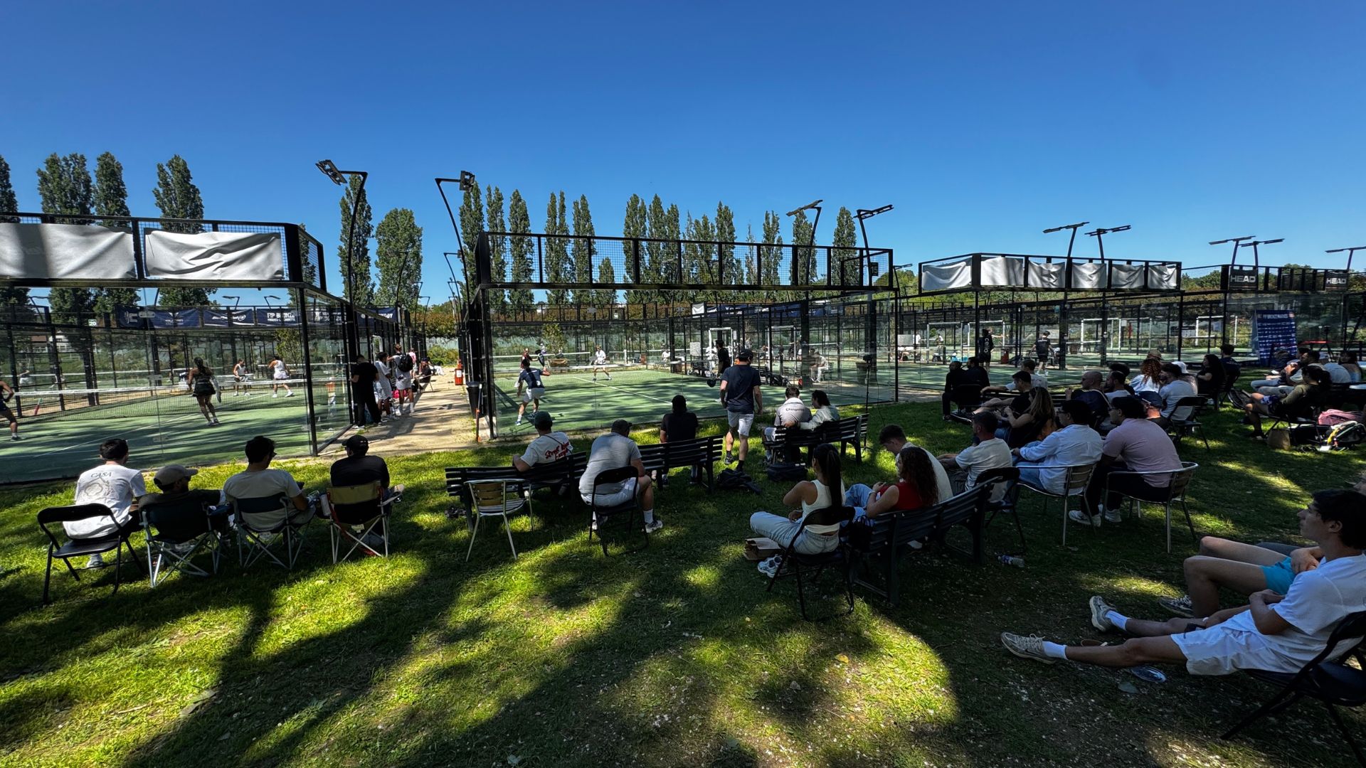 Crowd watching an outdoor padel match on fenced courts under a bright blue sky, with players in action and spectators on benches nearby.