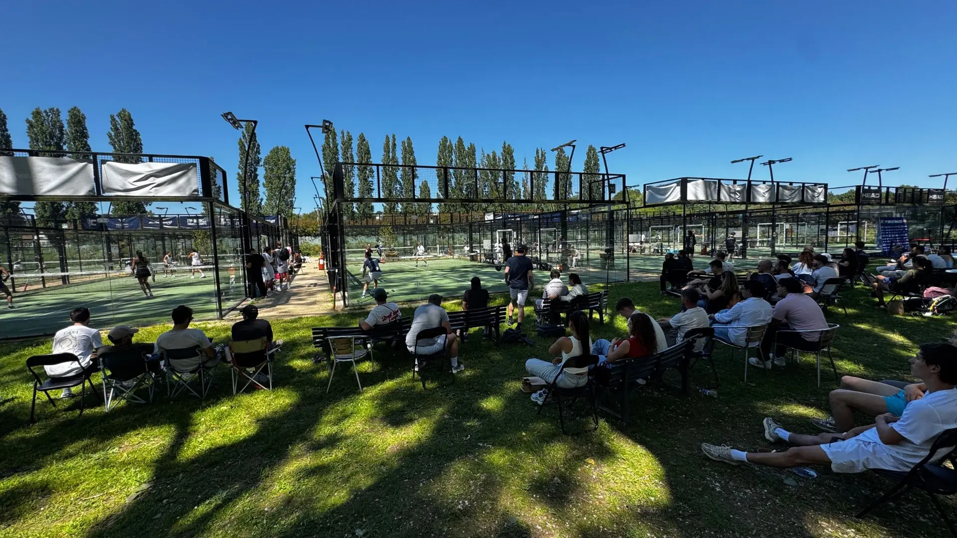 Crowd watching an outdoor padel match on fenced courts under a bright blue sky, with players in action and spectators on benches nearby.