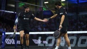 Two padel players in black outfits shake hands at the net after a match on an indoor court, paddles in hand.