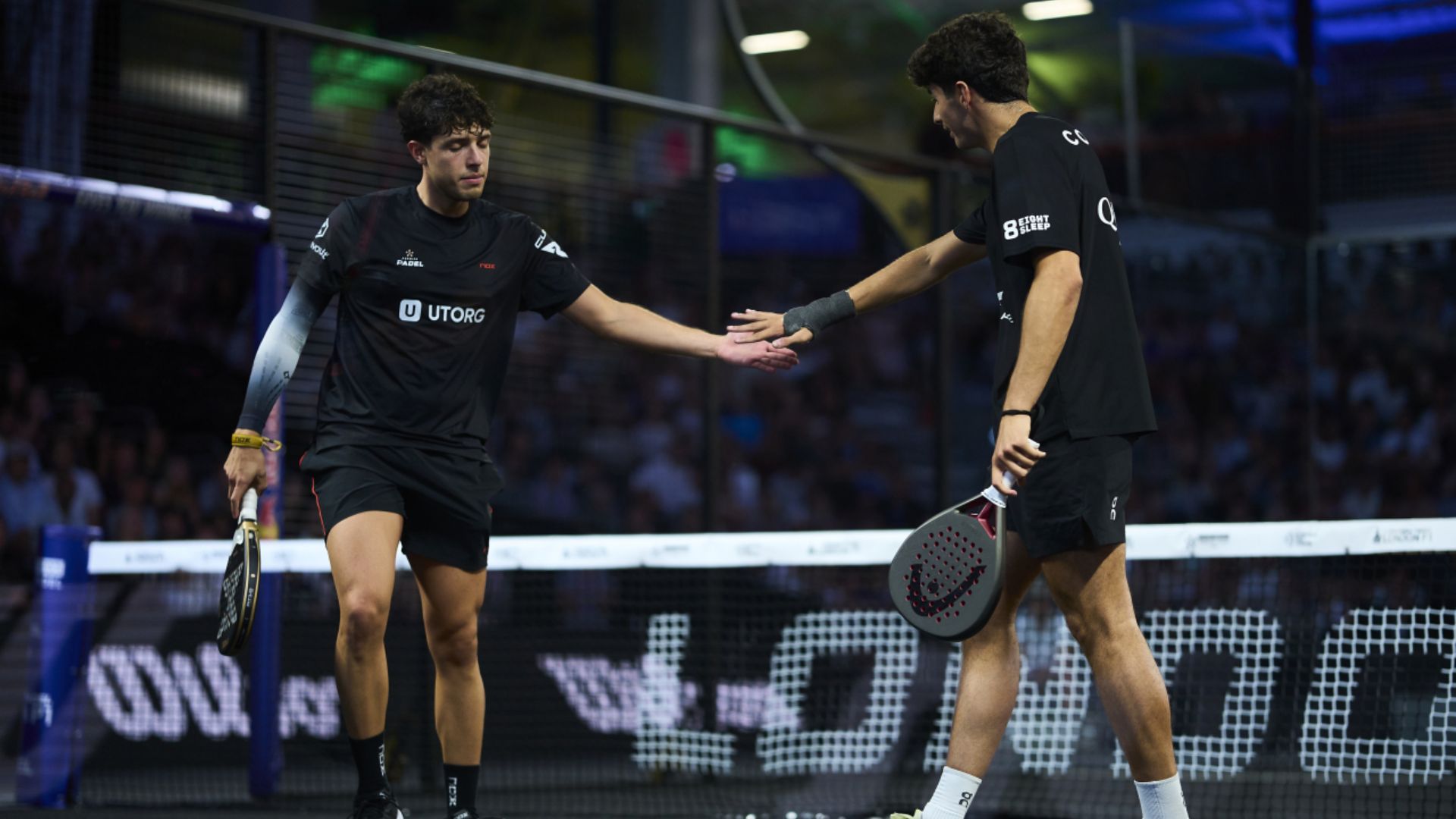 Two padel players in black outfits shake hands at the net after a match on an indoor court, paddles in hand.