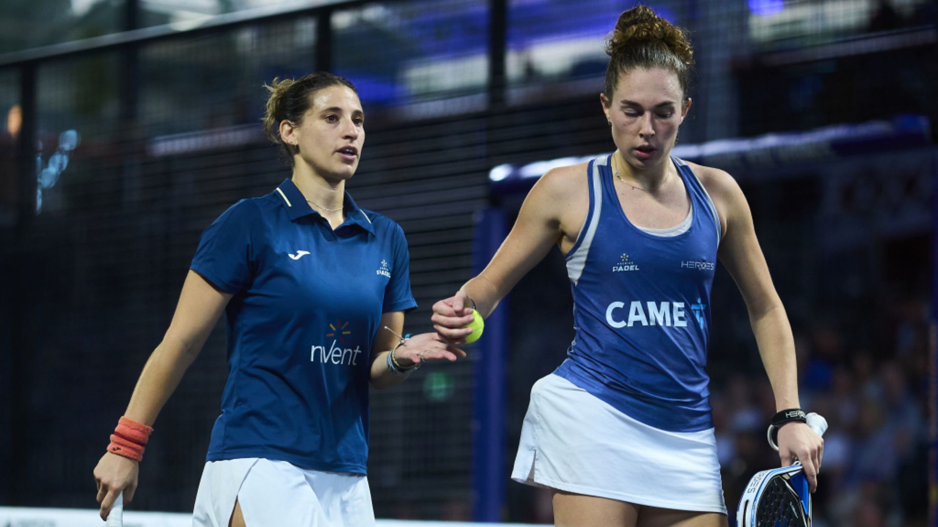 Two female padel players in blue shirts shake hands on the court after a match.