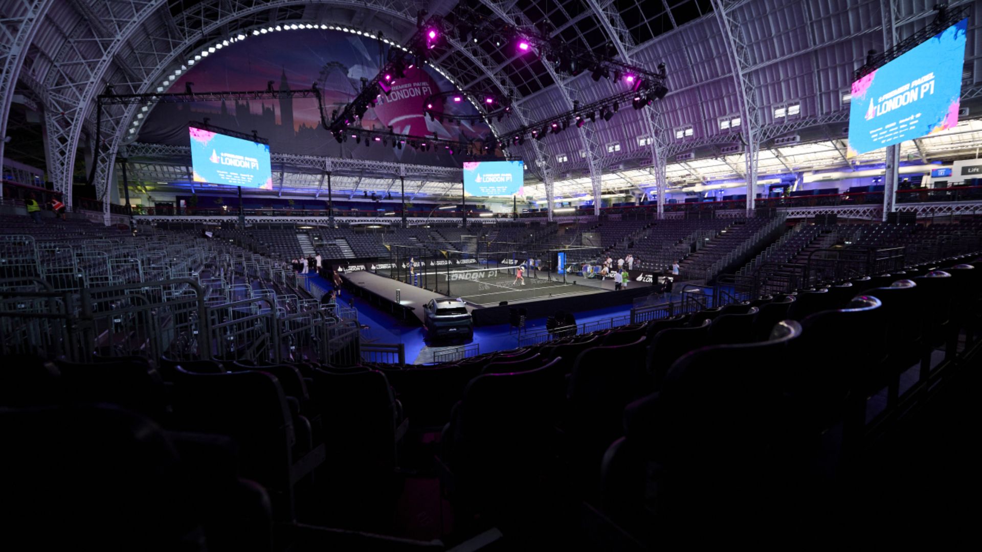 Indoor arena with a central competition court and surrounding empty seats, large screens display London PT banners under purple stage lights.