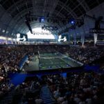Indoor tennis arena filled with spectators watching a match on a central court under bright overhead rigging and screens above.