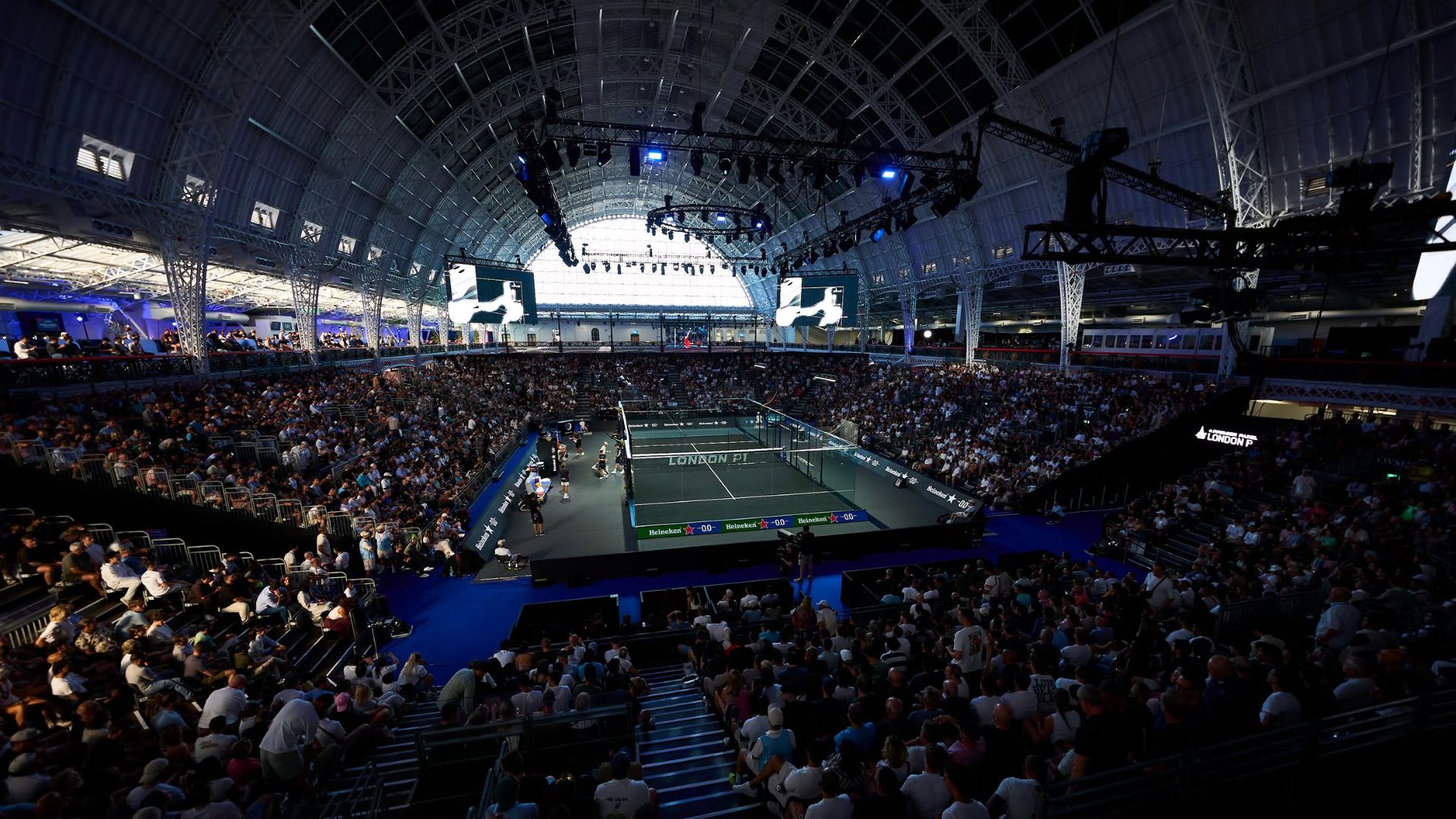 Indoor tennis arena filled with spectators watching a match on a central court under bright overhead rigging and screens above.