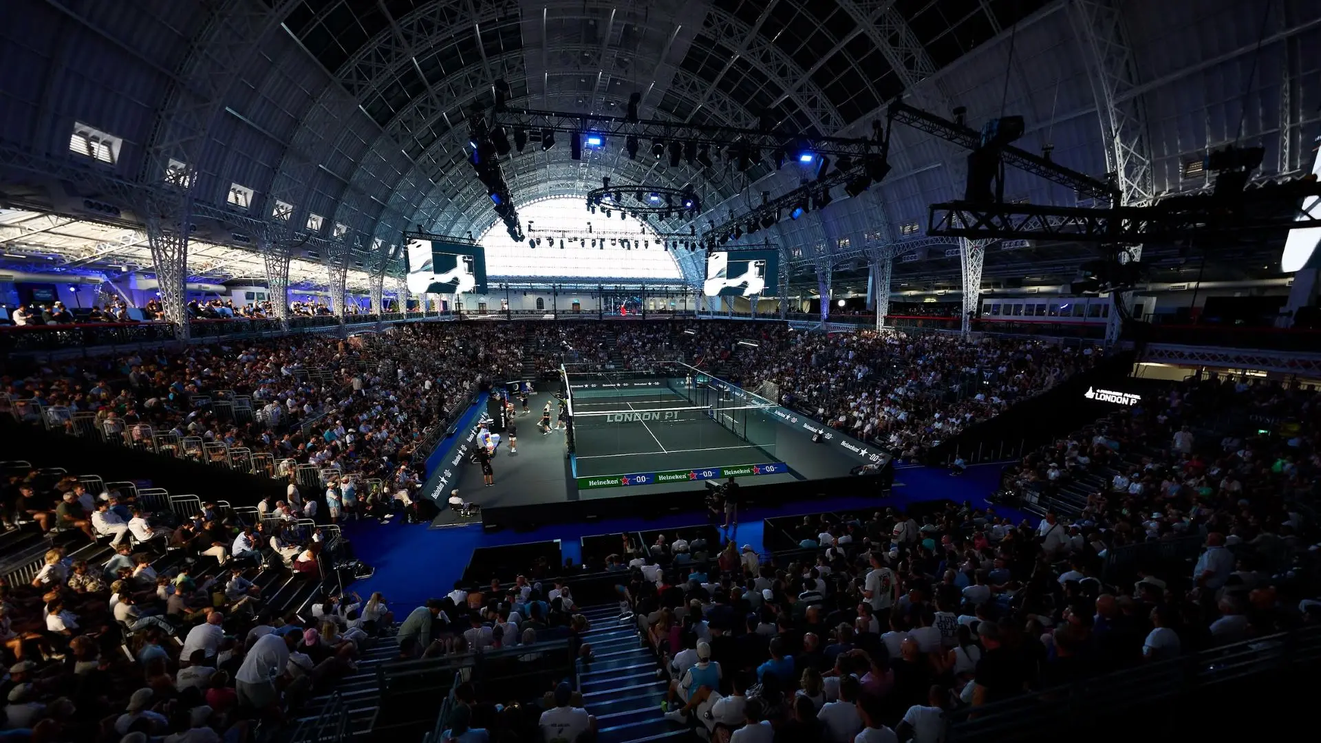 Indoor tennis arena filled with spectators watching a match on a central court under bright overhead rigging and screens above.
