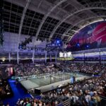 Indoor sports arena filled with spectators watching a padel match on a central court under a large curved steel roof and bright arena lighting. A scoreboard shows players Tapia/Coello and Di Nenno/Navarro nearby. Large banners read London P1.