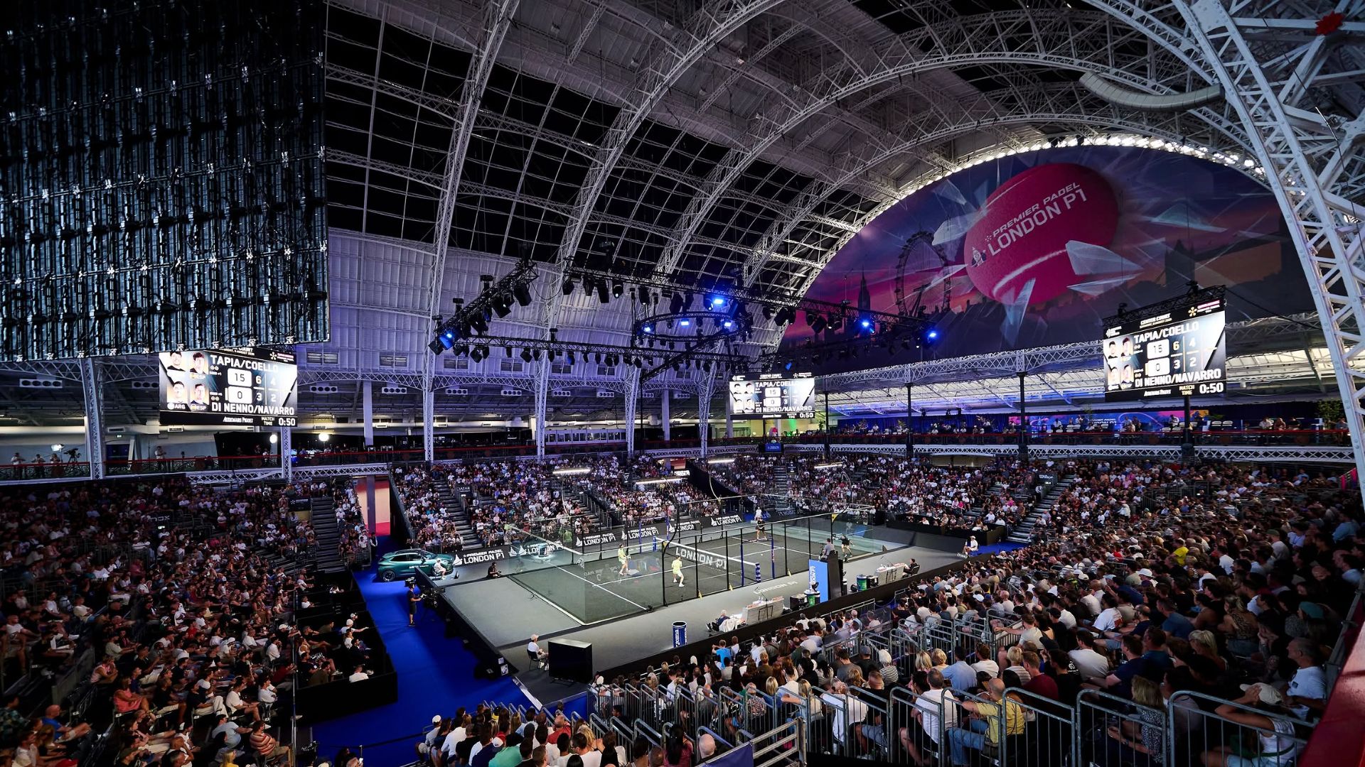 Indoor sports arena filled with spectators watching a padel match on a central court under a large curved steel roof and bright arena lighting. A scoreboard shows players Tapia/Coello and Di Nenno/Navarro nearby. Large banners read London P1.
