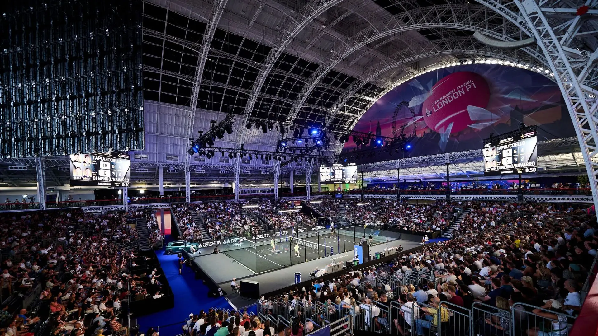 Indoor sports arena filled with spectators watching a padel match on a central court under a large curved steel roof and bright arena lighting. A scoreboard shows players Tapia/Coello and Di Nenno/Navarro nearby. Large banners read London P1.