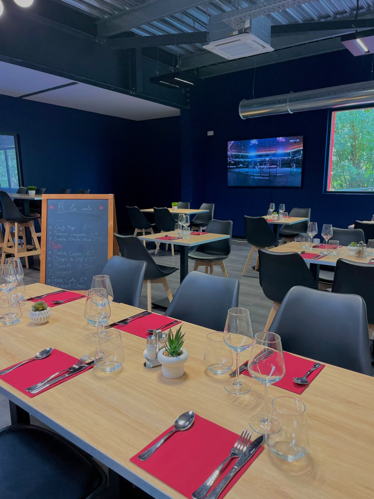 | Padel Magazine Light wood tables set with red placemats, glassware, and small potted plants in a modern blue-walled restaurant.