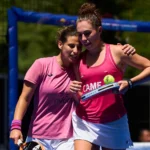 Two female tennis players in pink shirts celebrate together on a court, one holding a racket with a tennis ball tucked on the strings in celebration moment.