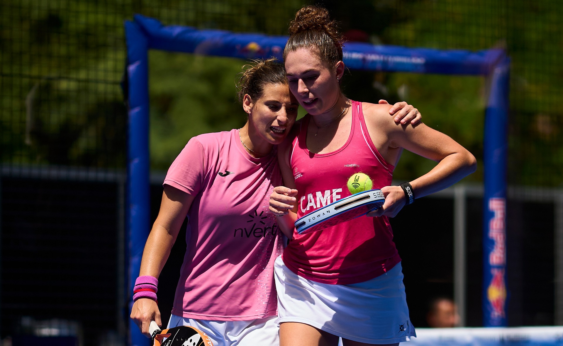 Two female tennis players in pink shirts celebrate together on a court, one holding a racket with a tennis ball tucked on the strings in celebration moment.