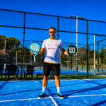 Man on a blue padel court holding two rackets, wearing sunglasses and a white logoed shirt, with fencing and trees behind him.