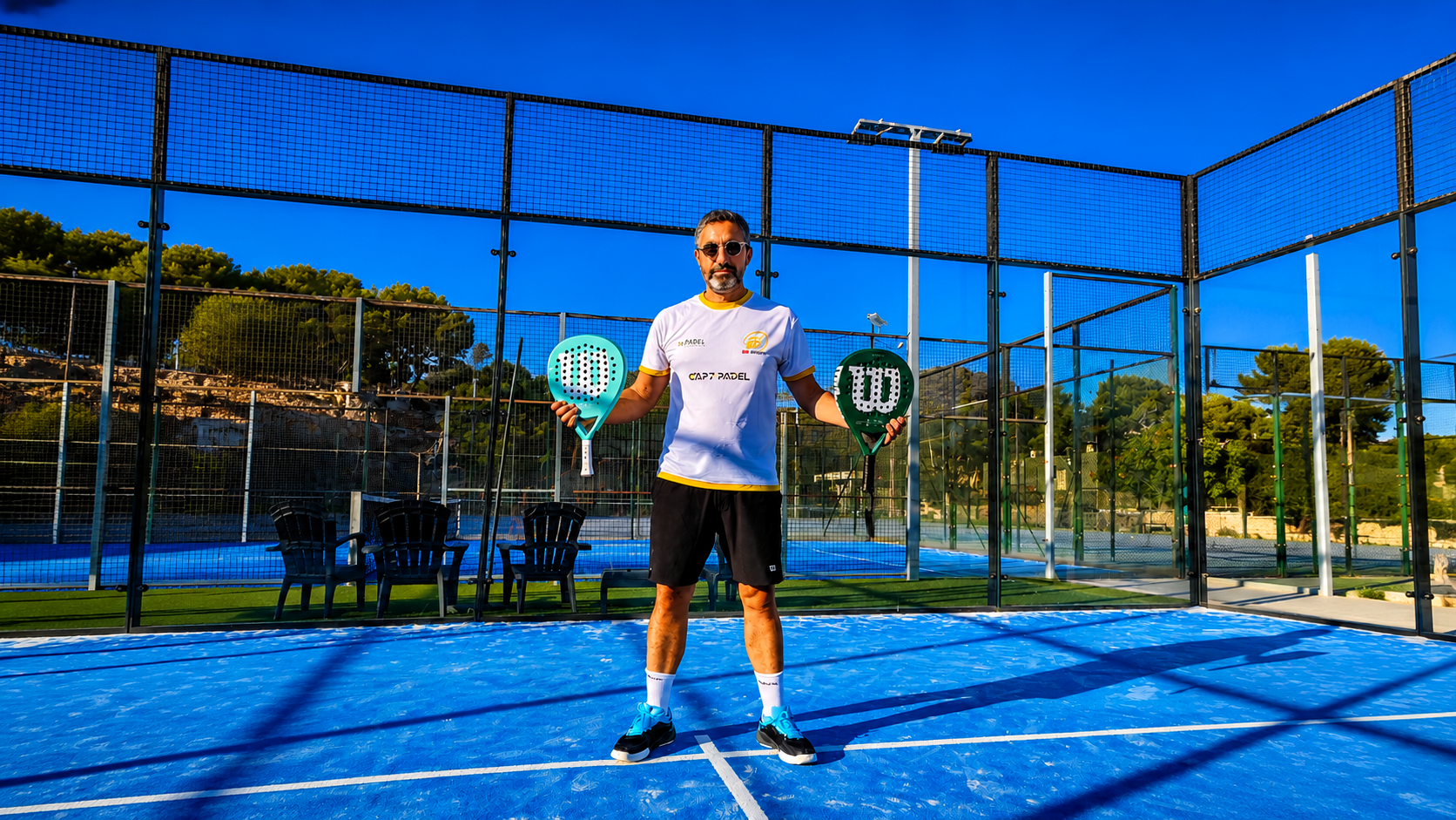 Man on a blue padel court holding two rackets, wearing sunglasses and a white logoed shirt, with fencing and trees behind him.
