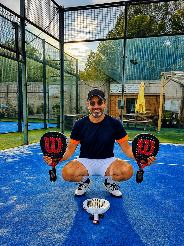 Stef Cassis 1 | Padel Magazine Man crouching on a blue padel court, holding two red Wilson padel rackets, wearing a navy shirt and white shorts, cap and sunglasses.